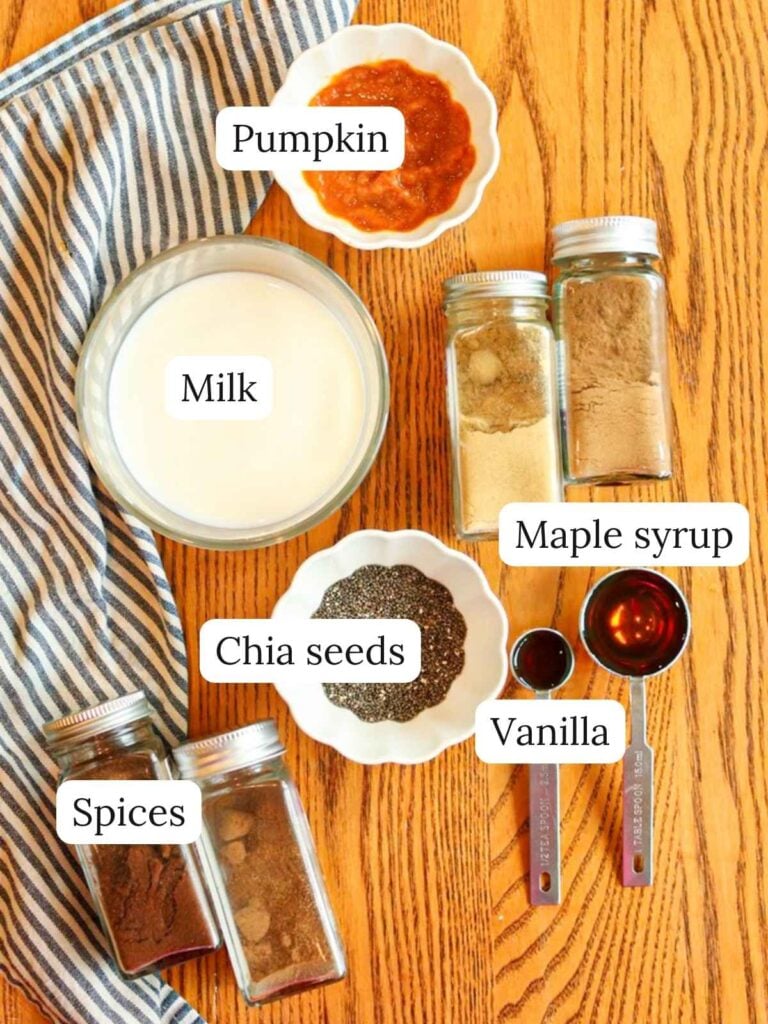 Labeled ingredients on a wooden table with a striped cloth napkin including milk, pumpkin, spices, maple syrup, vanilla, and chia seeds.
