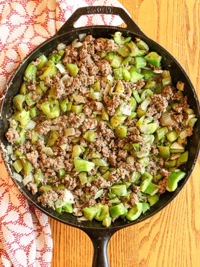 Browned ground beef, peppers, and onions in a skillet on a wooden surface.