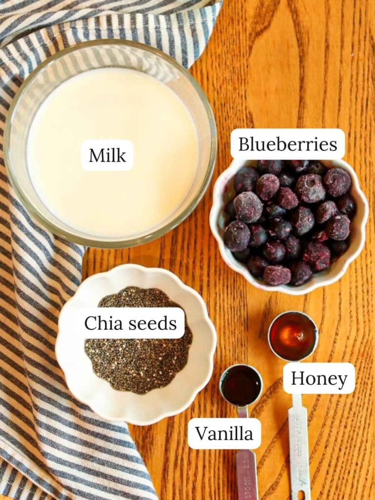 Labeled ingredients on a wood table with a cloth napkin including milk, chia seeds, blueberries, honey, and vanilla.