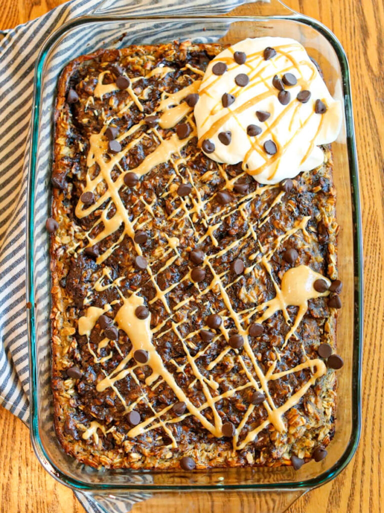 Chocolate baked oatmeal on a kitchen table topped with whipped cream, chocolate chips, and drizzled peanut butter.