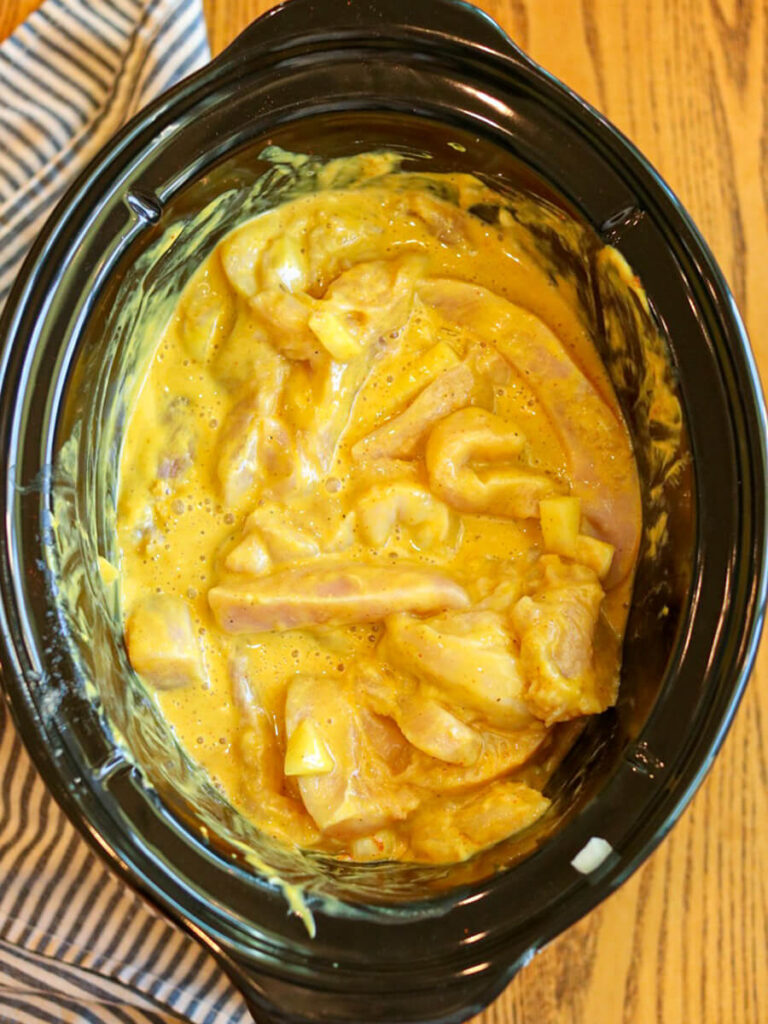 A slow cooker with raw chicken strips and a honey mustard sauce on a wooden surface.