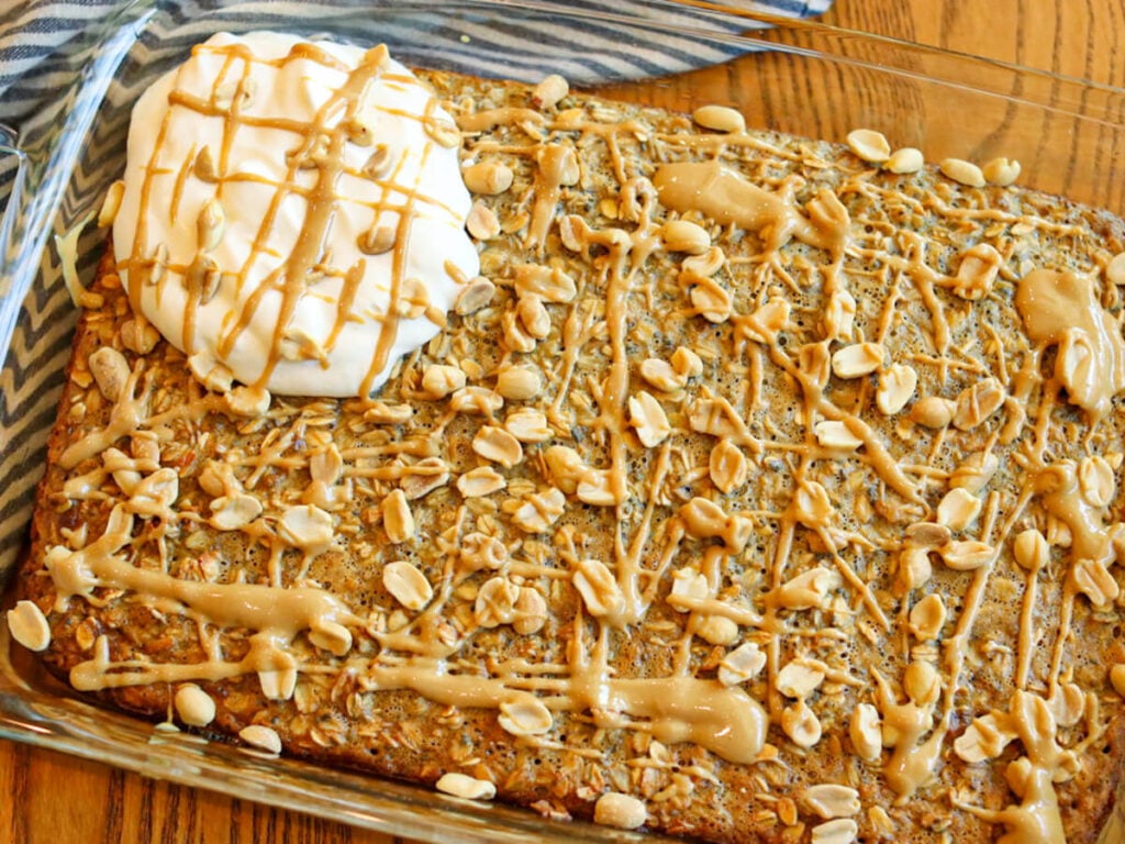 Peanut butter baked oatmeal drizzled with peanut butter and topped with roasted peanuts and whipped cream on a wood table.