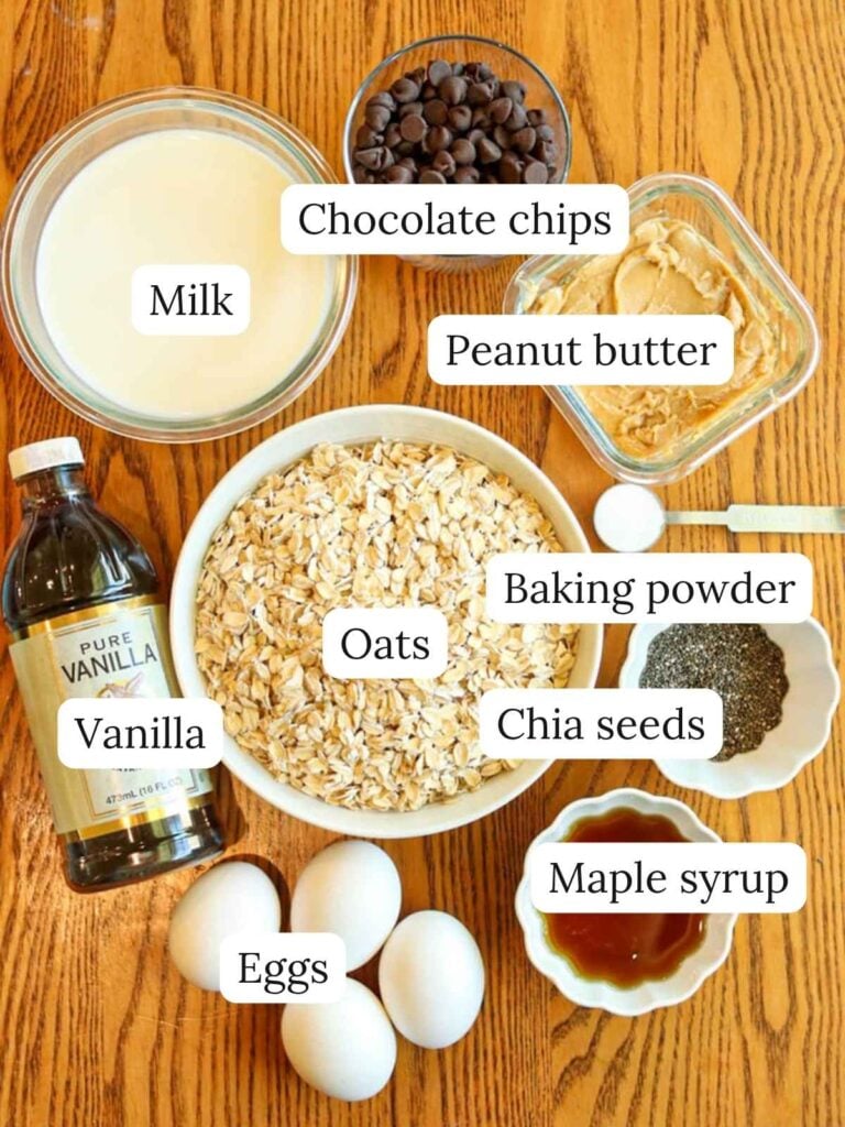 Labeled ingredients o a wood table including oats, peanut butter, maple syrup, vanilla, eggs, chia seeds, chocolate chips, milk, and baking powder.