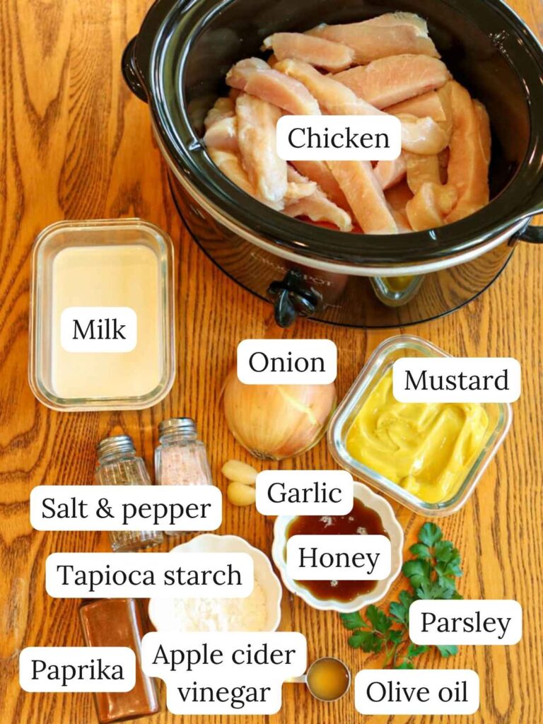 Labeled ingredients on a wooden table with a slow cooker including chicken, mustard, honey, milk, salt, pepper, paprika, parsley, garlic, onion, tapioca starch, apple cider vinegar, and olive oil.
