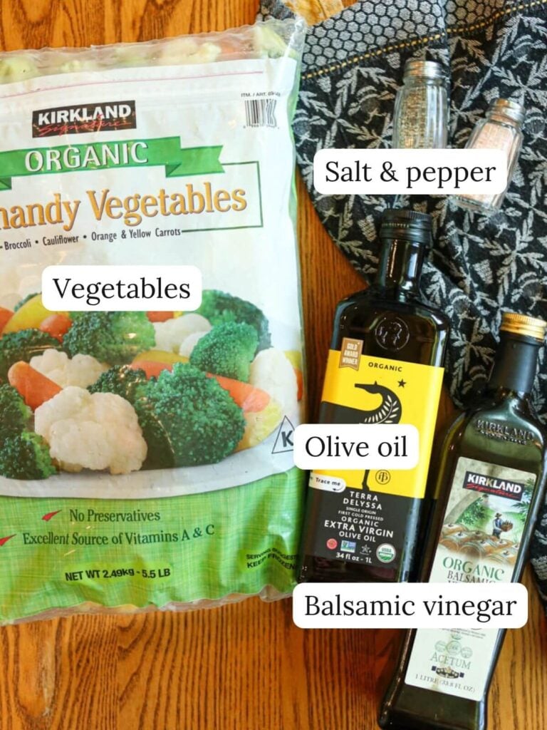 Labeled ingredients on a wood table with a navy kitchen towel including vegetables, salt and pepper, olive oil, and balsamic vinegar.