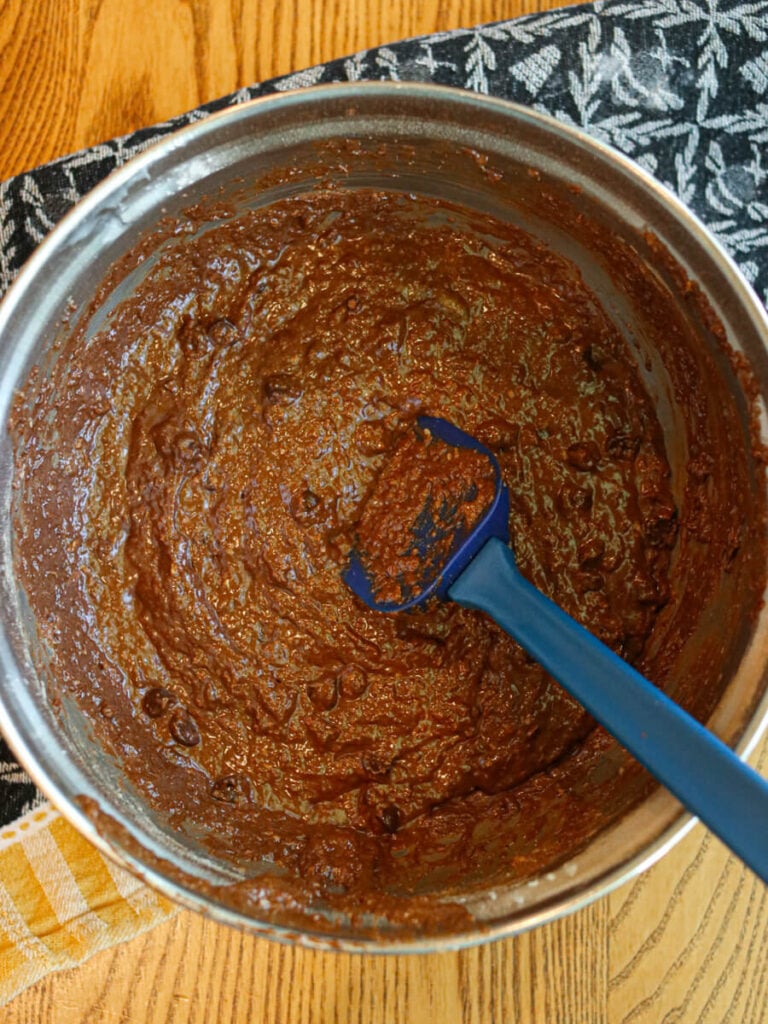 Gluten, egg, and dairy-free brownie batter in a mixing bowl on a wood table with a kitchen towel.
