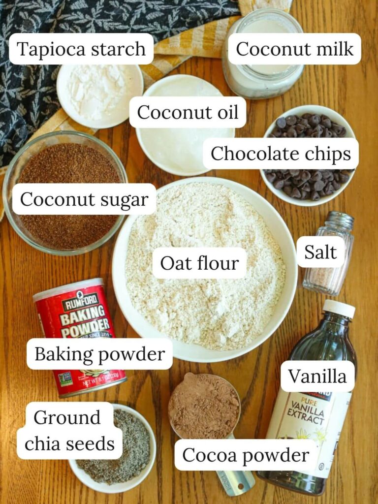 Labeled ingredients on a wood table including tapioca starch, coconut milk, coconut oil, chocolate chips, coconut sugar, oat flour, baking powder, salt, chia seeds, cocoa powder, and vanilla.