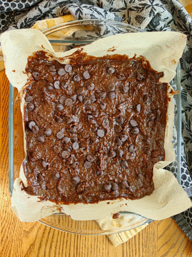 Oat flour brownie batter in a glass baking dish lined with parchment paper on a table.