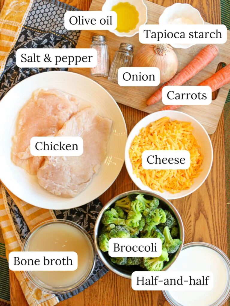 Labeled ingredients on a table including chicken, bone broth, broccoli, half-and-half, cheese, carrots, onion, salt and pepper, olive oil, and tapioca starch.