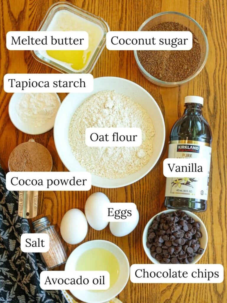 Labeled ingredients on a kitchen table including melted butter, cocoa powder, oat flour, eggs, chocolate chips, and avocado oil